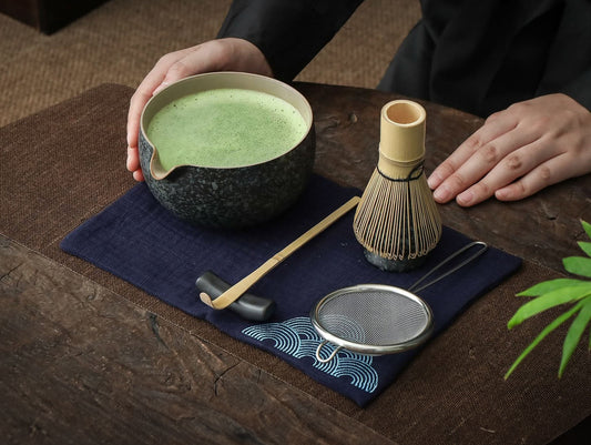 A person's hands holding a ceramic bowl of matcha, alongside a bamboo whisk, ceramic scoop stand, and a matcha powder sifter, all arranged on a dark cloth.