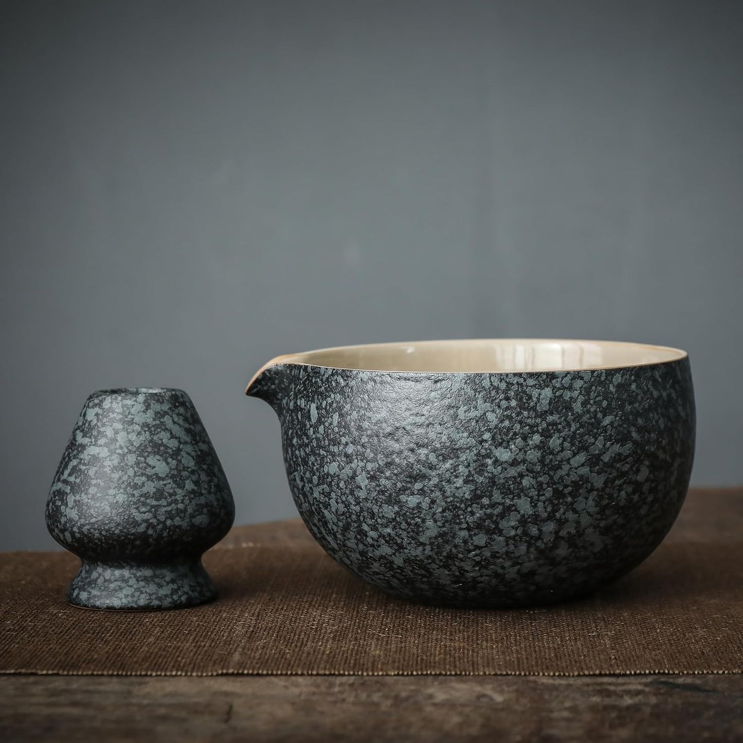 A person's hands holding a ceramic bowl of matcha, alongside a bamboo whisk, ceramic scoop stand, and a matcha powder sifter, all arranged on a dark cloth.
