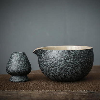 A person's hands holding a ceramic bowl of matcha, alongside a bamboo whisk, ceramic scoop stand, and a matcha powder sifter, all arranged on a dark cloth.