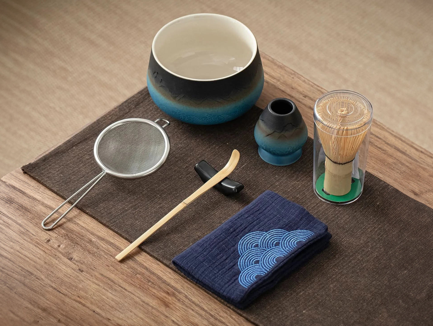 A set of matcha tea ceremony equipment laid out on a wooden table, including a ceramic bowl, bamboo whisk, bamboo scoop, ceramic whisk holder, ceramic scoop stand, cotton tea cloth, and a matcha powder sifter.