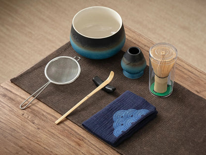 A set of matcha tea ceremony equipment laid out on a wooden table, including a ceramic bowl, bamboo whisk, bamboo scoop, ceramic whisk holder, ceramic scoop stand, cotton tea cloth, and a matcha powder sifter.