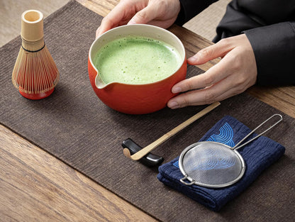 A person is preparing matcha tea with a red ceramic bowl, a bamboo whisk, and a blue-patterned ceramic scoop stand on a table. There is also a blue cotton tea cloth and a matcha powder sifter present.
