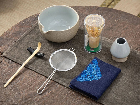 A set of matcha ceremony tools including a bamboo whisk, a bamboo scoop, a ceramic bowl, a ceramic whisk holder, a ceramic scoop stand, a cotton tea cloth, and a matcha powder sifter, displayed on a wooden surface.