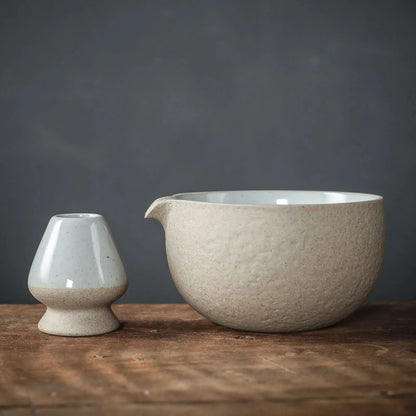 A set of matcha ceremony tools including a bamboo whisk, a bamboo scoop, a ceramic bowl, a ceramic whisk holder, a ceramic scoop stand, a cotton tea cloth, and a matcha powder sifter, displayed on a wooden surface.