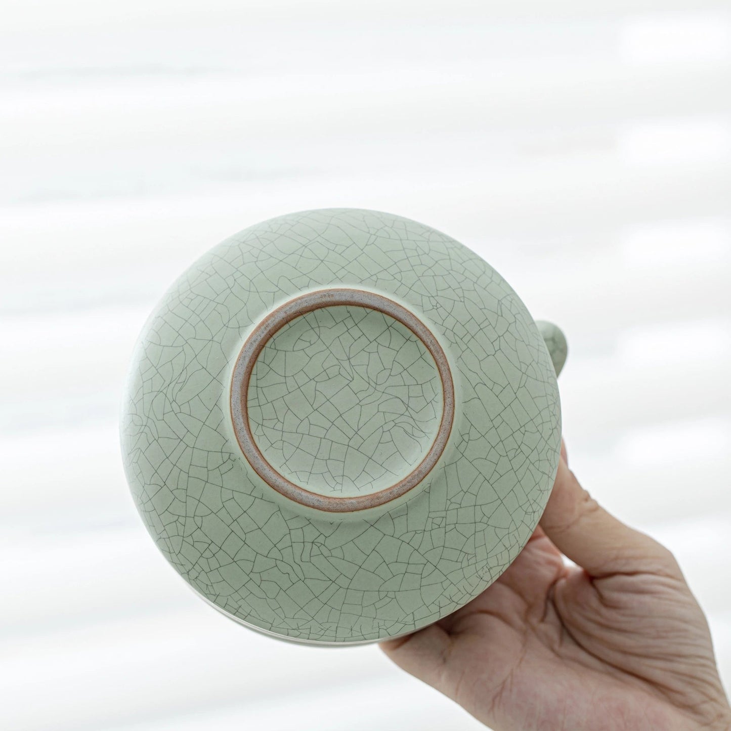 Light green ceramic teapot with a cracked glaze pattern held by a hand against a white background