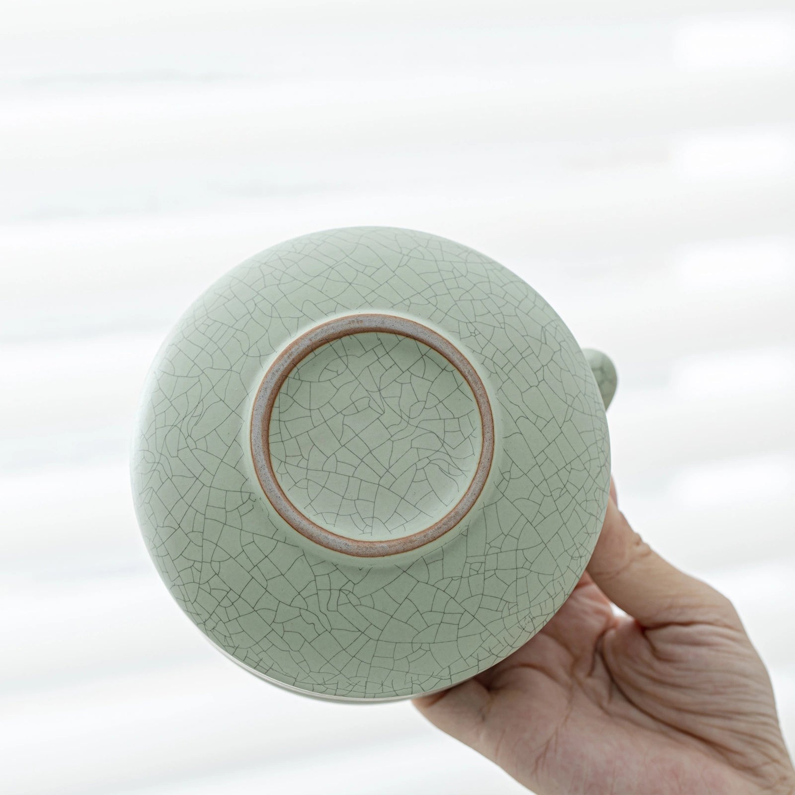Light green ceramic teapot with a cracked glaze pattern held by a hand against a white background