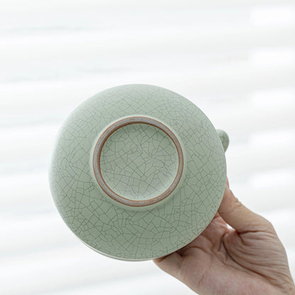 Light green ceramic teapot with a cracked glaze pattern held by a hand against a white background
