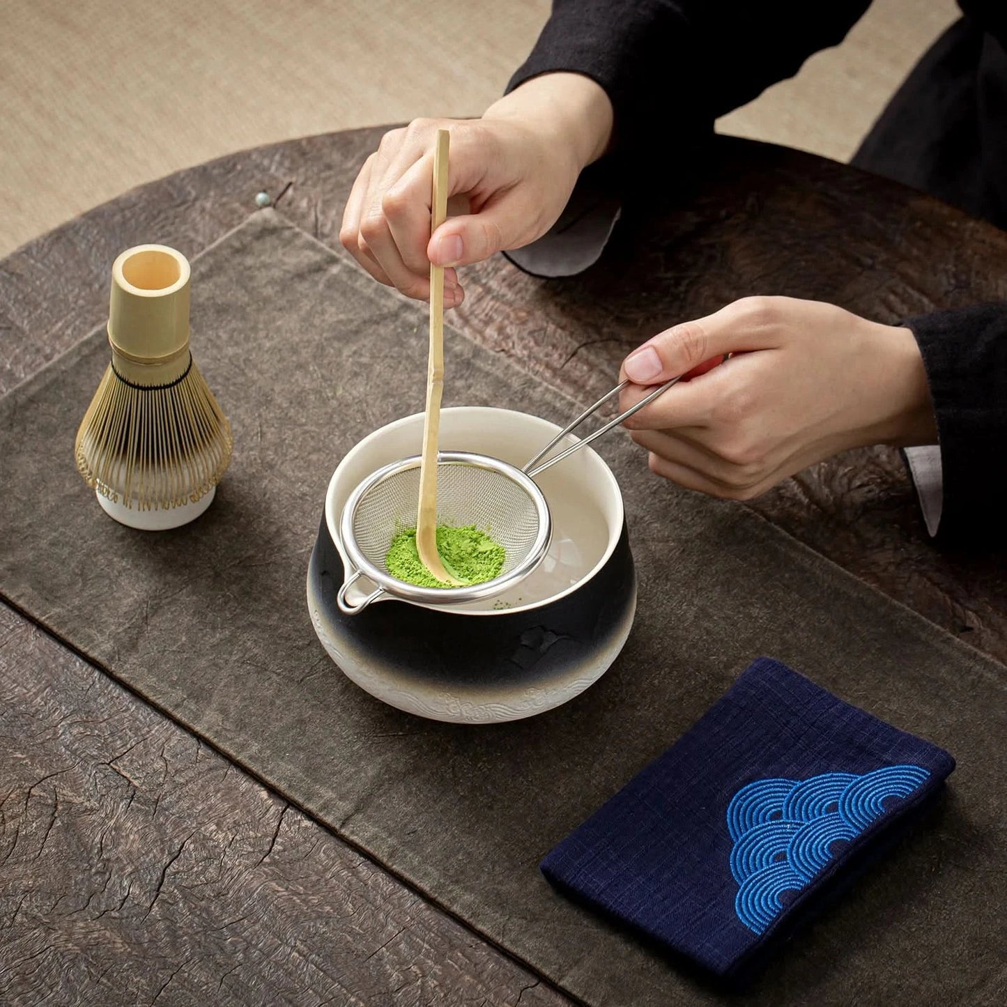 Person preparing matcha tea with a whisk and a blue cloth on a wooden table.
