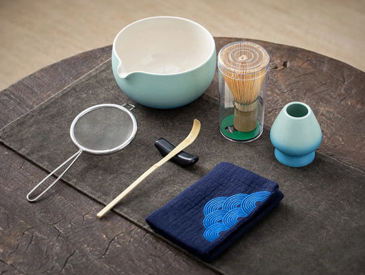 A set of matcha tea ceremony equipment laid out on a wooden surface, including a ceramic bowl, bamboo whisk, bamboo scoop, ceramic whisk holder, ceramic scoop stand, cotton tea cloth, and a matcha powder sifter, with a blue lagoon color theme.