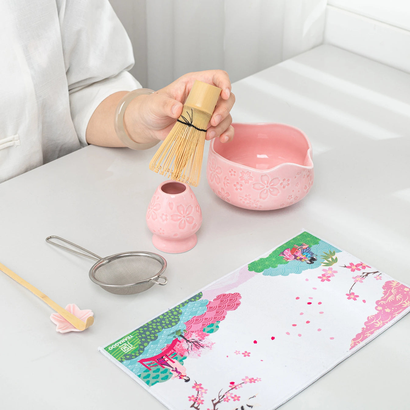 Person holding a bamboo whisk over a pink bowl with tea-making tools on a table.