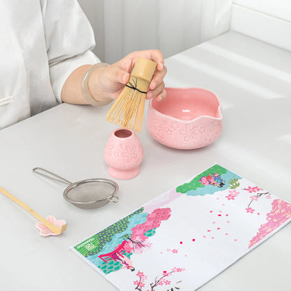 Person holding a bamboo whisk over a pink bowl with tea-making tools on a table.