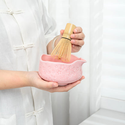 Person holding a pink ceramic whisk and bowl with a wooden whisk against a white background