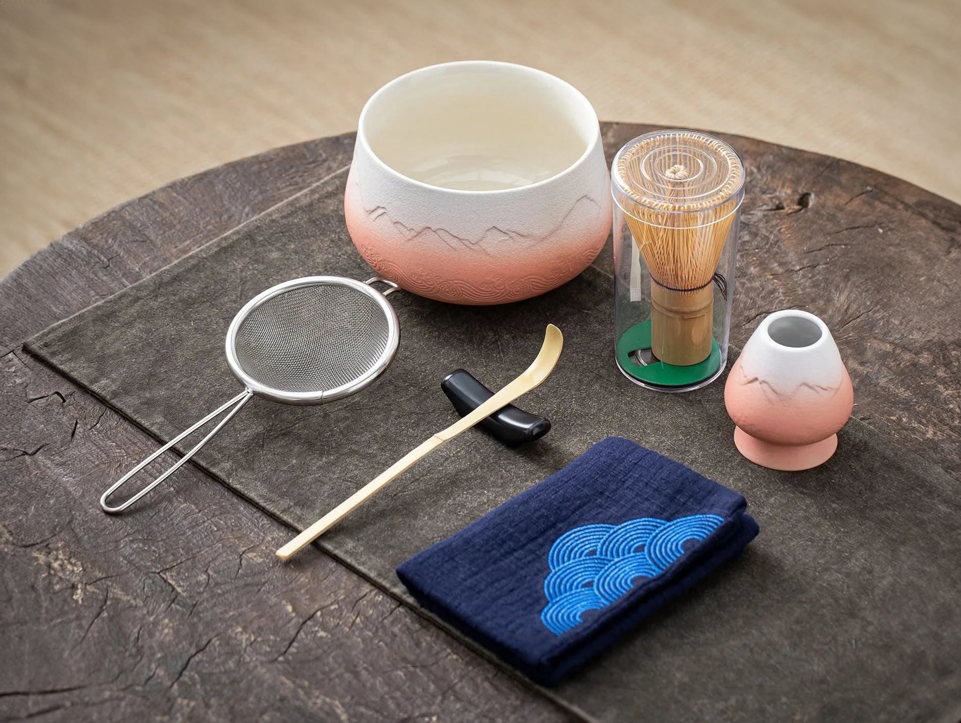 A set of matcha ceremony tools laid out on a wooden surface, including a ceramic bowl, bamboo whisk, bamboo scoop, ceramic whisk holder, ceramic scoop stand, cotton tea cloth, and a matcha powder sifter, with a pink and blue textured background.