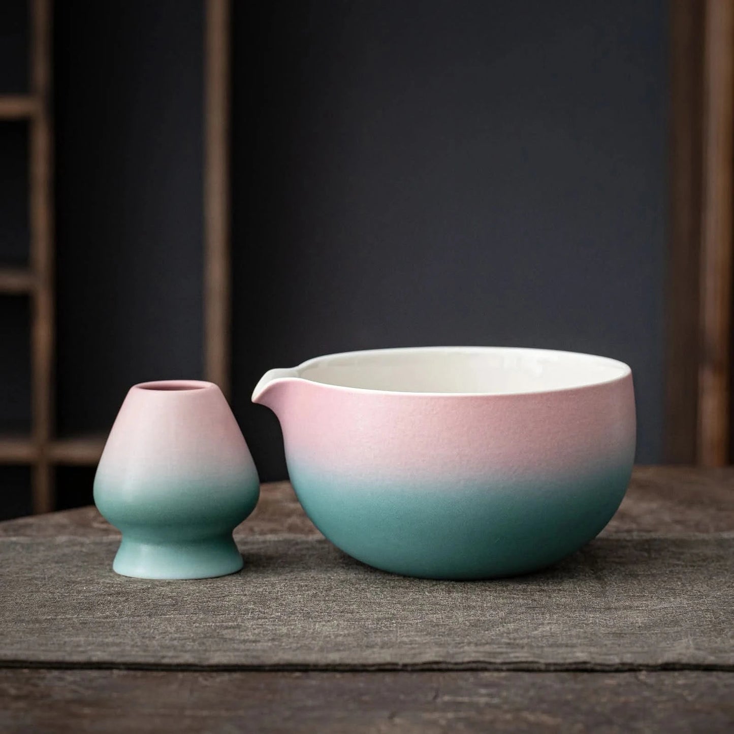 A set of matcha tea ceremony equipment including a pink and blue ceramic bowl, a bamboo whisk, a bamboo scoop, a ceramic whisk holder, a ceramic scoop stand, a cotton tea cloth, and a matcha powder sifter, displayed on a wooden surface.