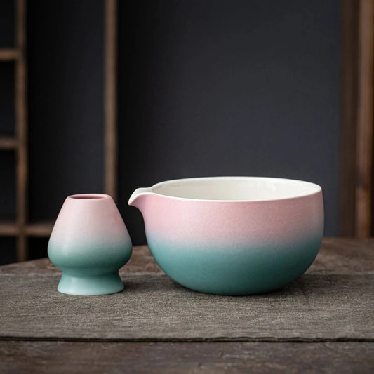 A set of matcha tea ceremony equipment including a pink and blue ceramic bowl, a bamboo whisk, a bamboo scoop, a ceramic whisk holder, a ceramic scoop stand, a cotton tea cloth, and a matcha powder sifter, displayed on a wooden surface.
