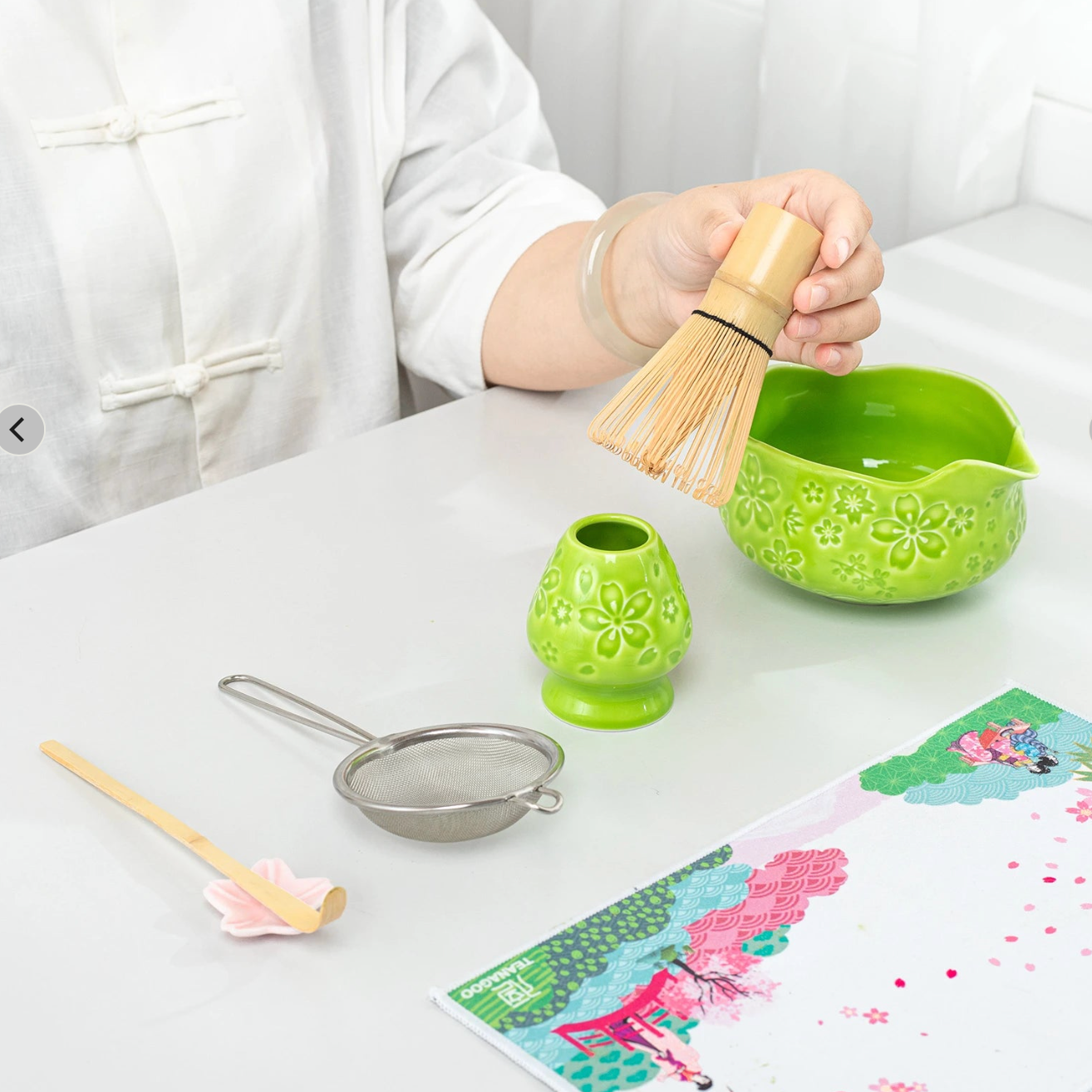 Person preparing tea with a green bowl, whisk, and other utensils on a table.