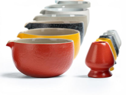 A person is preparing matcha tea with a red ceramic bowl, a bamboo whisk, and a blue-patterned ceramic scoop stand on a table. There is also a blue cotton tea cloth and a matcha powder sifter present.
